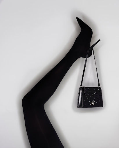 Black high-heel shoe and matching shoulder bag against a white background. Leg is raised in a bent position to display heel.