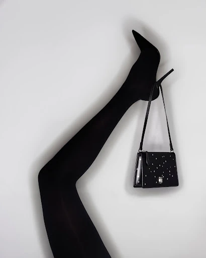 Black high-heel shoe and matching shoulder bag against a white background. Leg is raised in a bent position to display heel.
