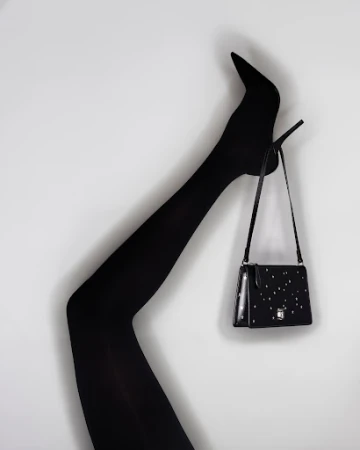 Black high-heel shoe and matching shoulder bag against a white background. Leg is raised in a bent position to display heel.