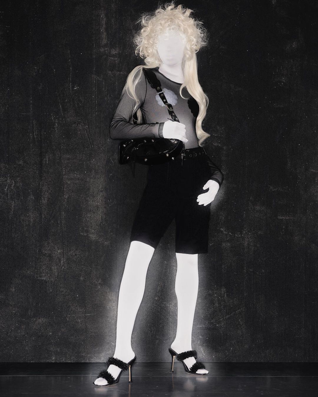 Full shot of a mannequin wearing a sheer top, black shorts, tights, and heeled sandals, posing against a dark textured backdrop.