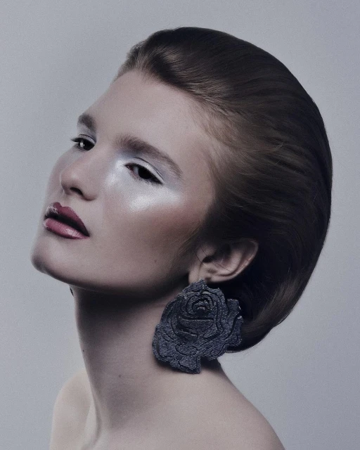 Close-up portrait of a woman with dramatic white eyeshadow and dark pink lipstick, wearing large rose-shaped denim earrings.