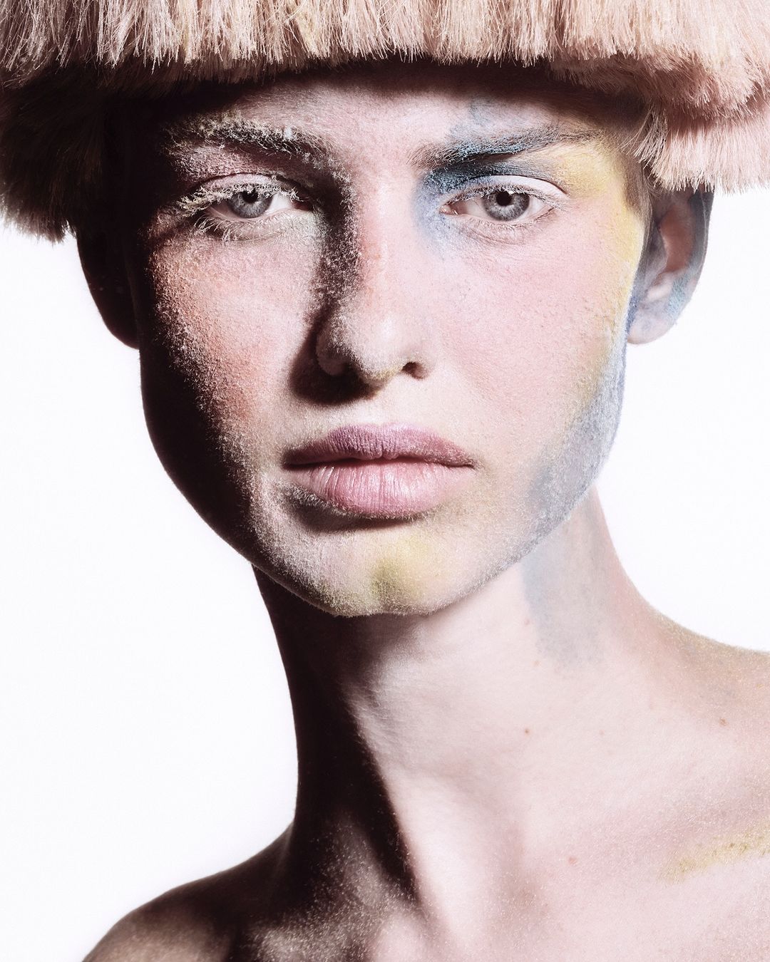 Close-up portrait of a person with creative makeup in blue, yellow, and white tones, wearing a straw-like headpiece on a stark white background.