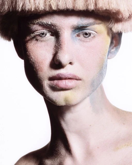 Close-up portrait of a person with creative makeup in blue, yellow, and white tones, wearing a straw-like headpiece on a stark white background.