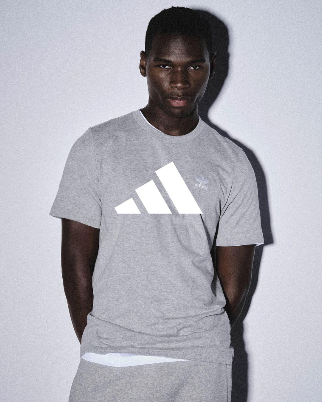 Dark-skinned adult male in a gray Adidas t-shirt and sweatpants posing indoors against a pale background in soft, diffused lighting.