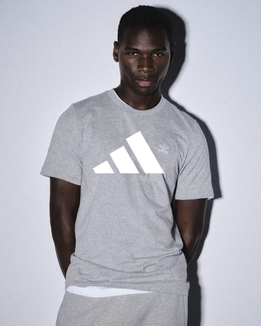 Dark-skinned adult male in a gray Adidas t-shirt and sweatpants posing indoors against a pale background in soft, diffused lighting.