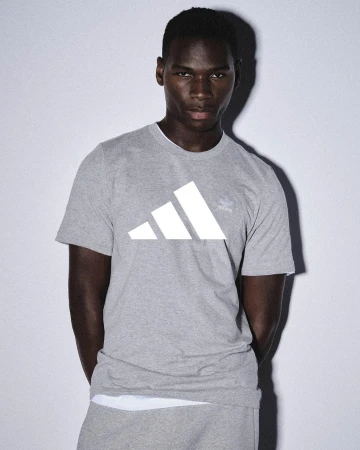 Dark-skinned adult male in a gray Adidas t-shirt and sweatpants posing indoors against a pale background in soft, diffused lighting.