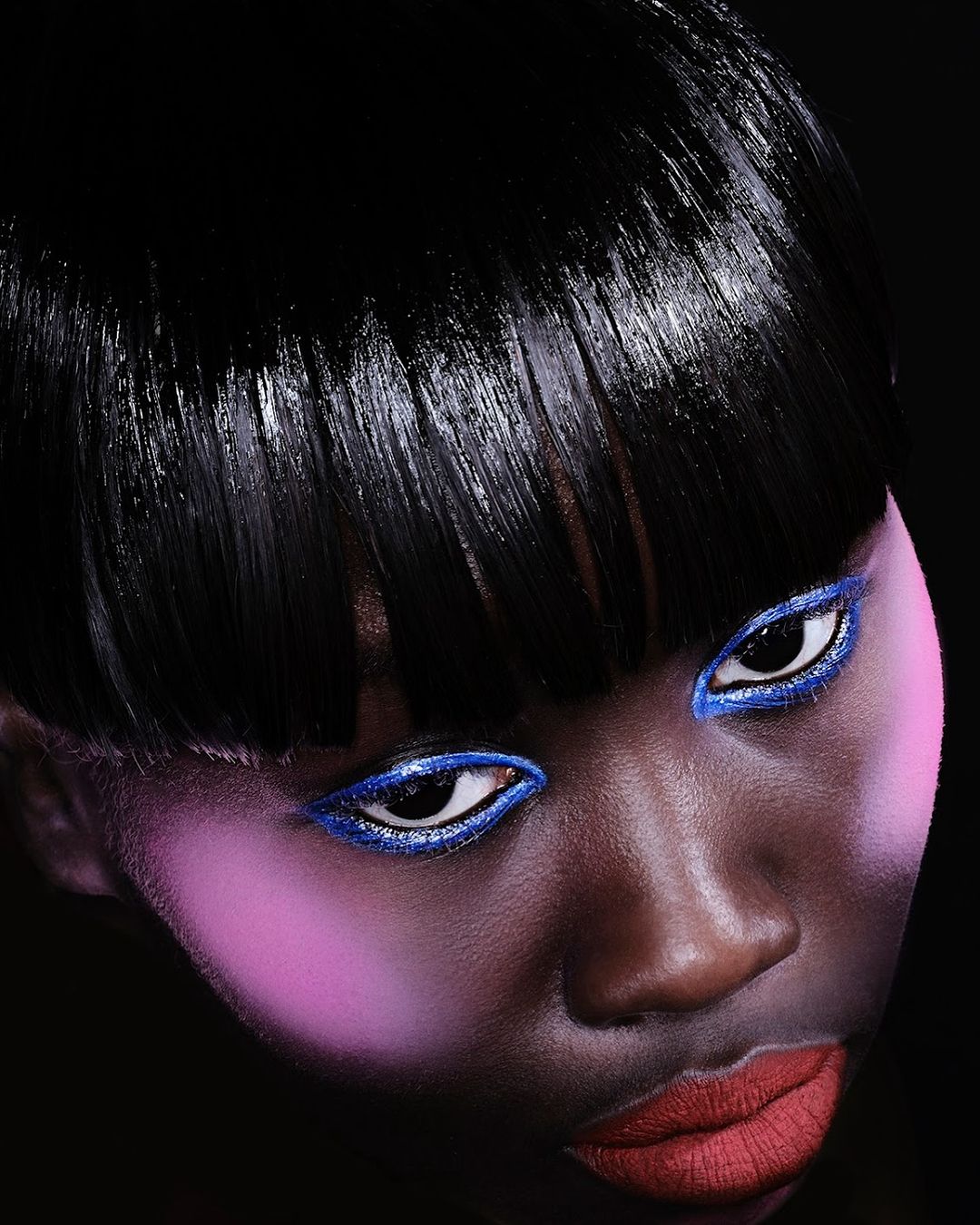 Close-up portrait of a woman with dark skin and glossy black hair, featuring bold blue eye makeup and pink blush on a dark background.