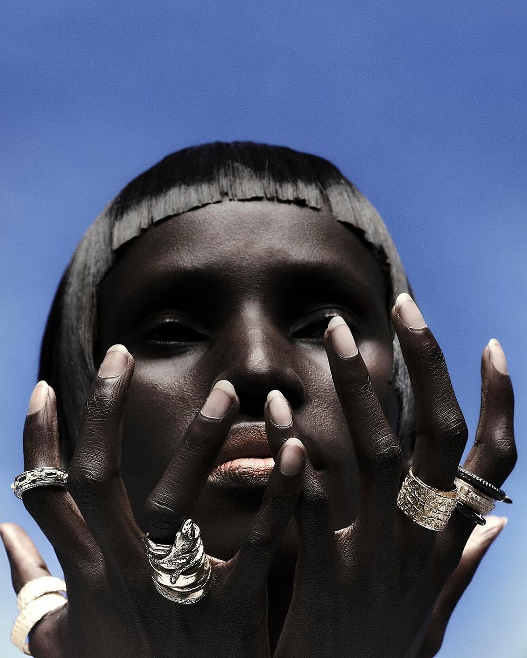 Close-up fashion shot of a dark-skinned woman with silver rings on each finger against a bright blue sky backdrop in outdoor lighting.