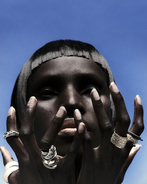 Close-up fashion shot of a dark-skinned woman with silver rings on each finger against a bright blue sky backdrop in outdoor lighting.