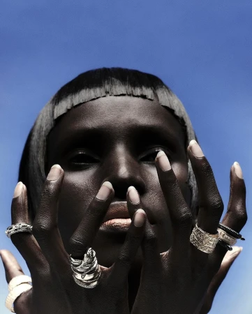 Close-up fashion shot of a dark-skinned woman with silver rings on each finger against a bright blue sky backdrop in outdoor lighting.