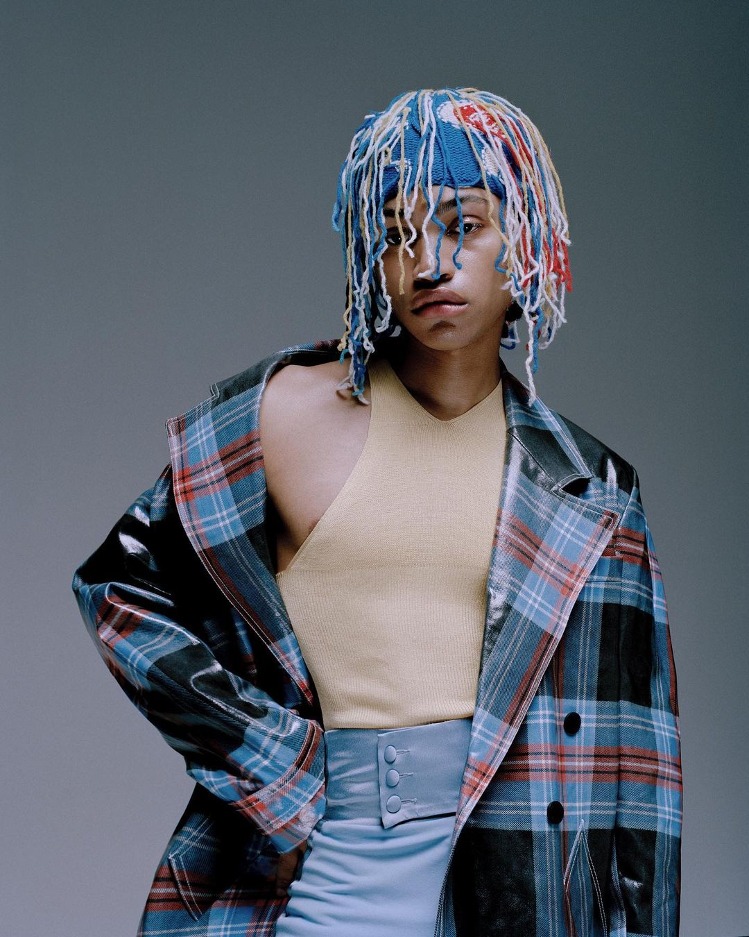 Person with colorful yarn hat, tan top, light blue skirt, and a plaid jacket posing in a photo studio with a light gray background.