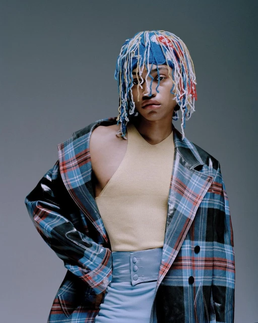 Person with colorful yarn hat, tan top, light blue skirt, and a plaid jacket posing in a photo studio with a light gray background.