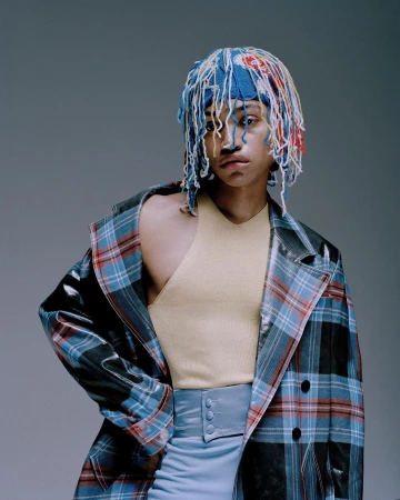 Person with colorful yarn hat, tan top, light blue skirt, and a plaid jacket posing in a photo studio with a light gray background.