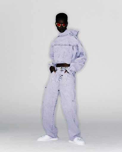 A person stands in a studio wearing a light purple denim outfit with a hooded crop-top jacket, loose jeans, sunglasses and white platform slides.