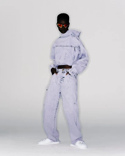 A person stands in a studio wearing a light purple denim outfit with a hooded crop-top jacket, loose jeans, sunglasses and white platform slides.