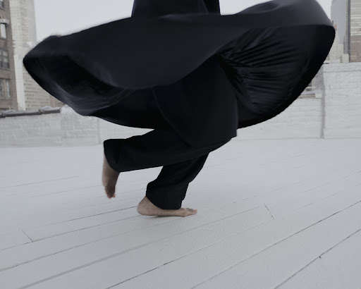 Barefoot person in black clothing is in motion on a white rooftop with cityscape buildings in the distance, captured in wide shot.