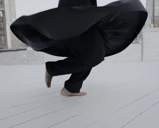Barefoot person in black clothing is in motion on a white rooftop with cityscape buildings in the distance, captured in wide shot.