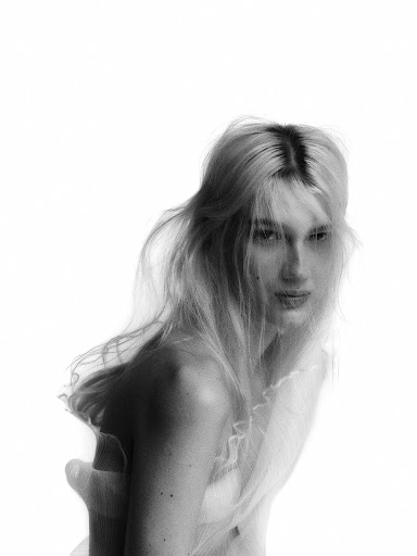 Black-and-white portrait of a blonde woman with fair skin, soft lighting, and a semi-transparent tulle fabric partially covering her.