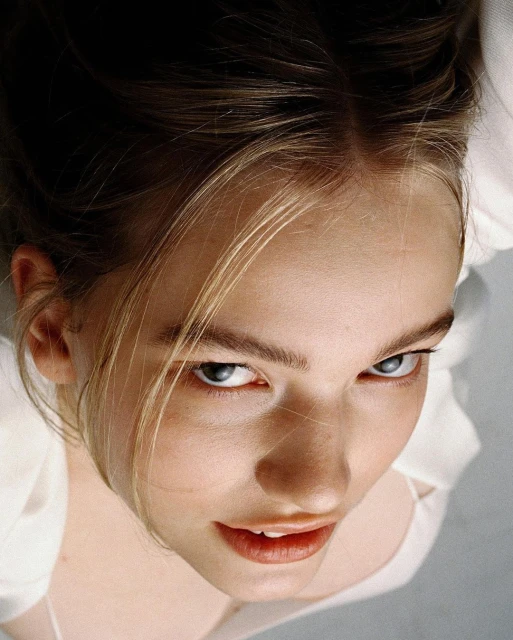Close-up, low-angle portrait of a fair-skinned young adult woman with blonde-streaked brown hair, looking upward with a neutral expression.