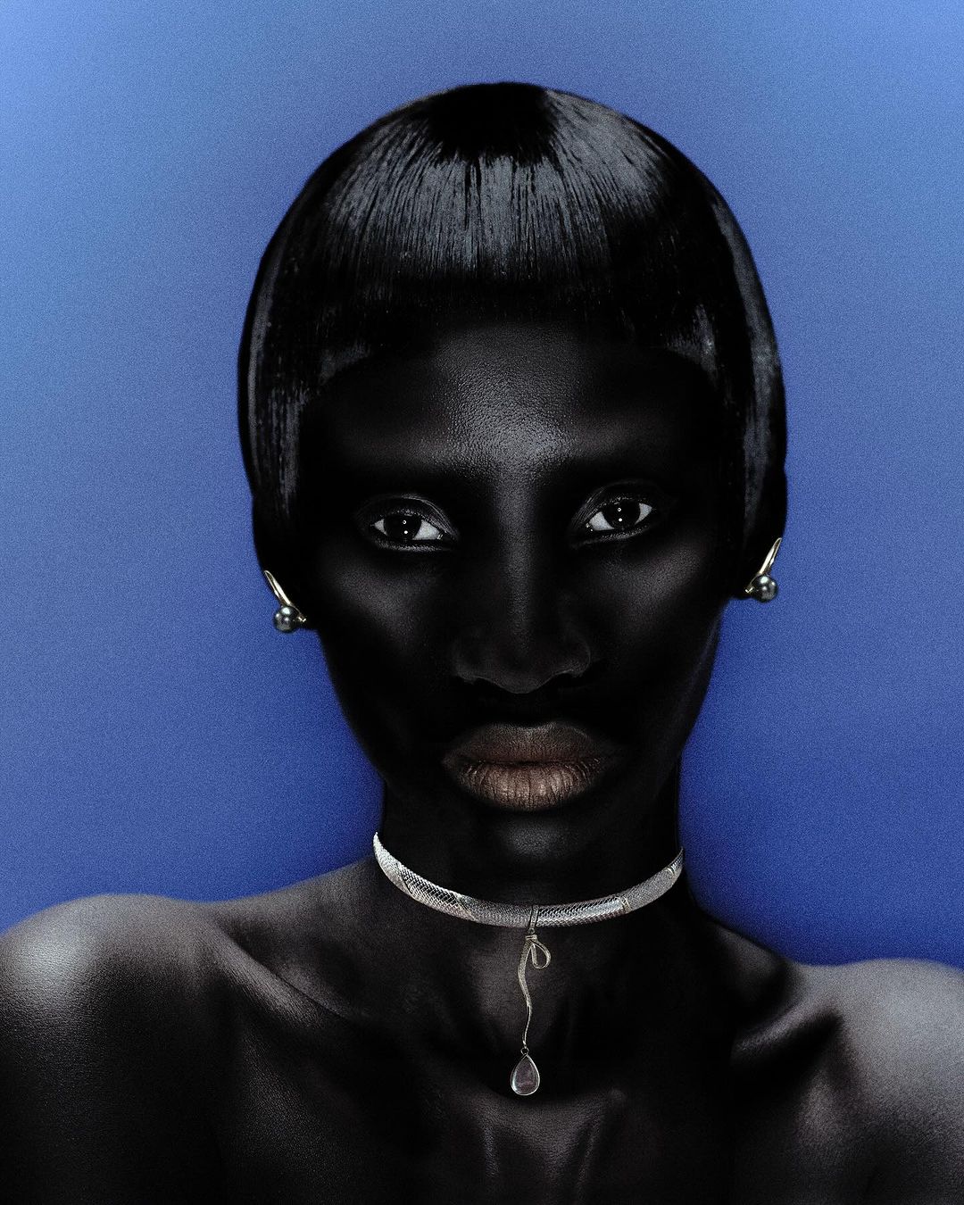 Close-up studio portrait of a dark-skinned woman with a short, shiny black hairstyle, wearing a silver choker against a blue background.