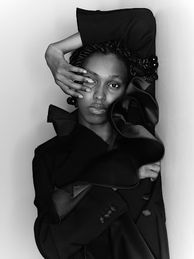 Monochrome fashion portrait of a Black woman in a coat, posing with a hand covering her eye, against a neutral studio backdrop.