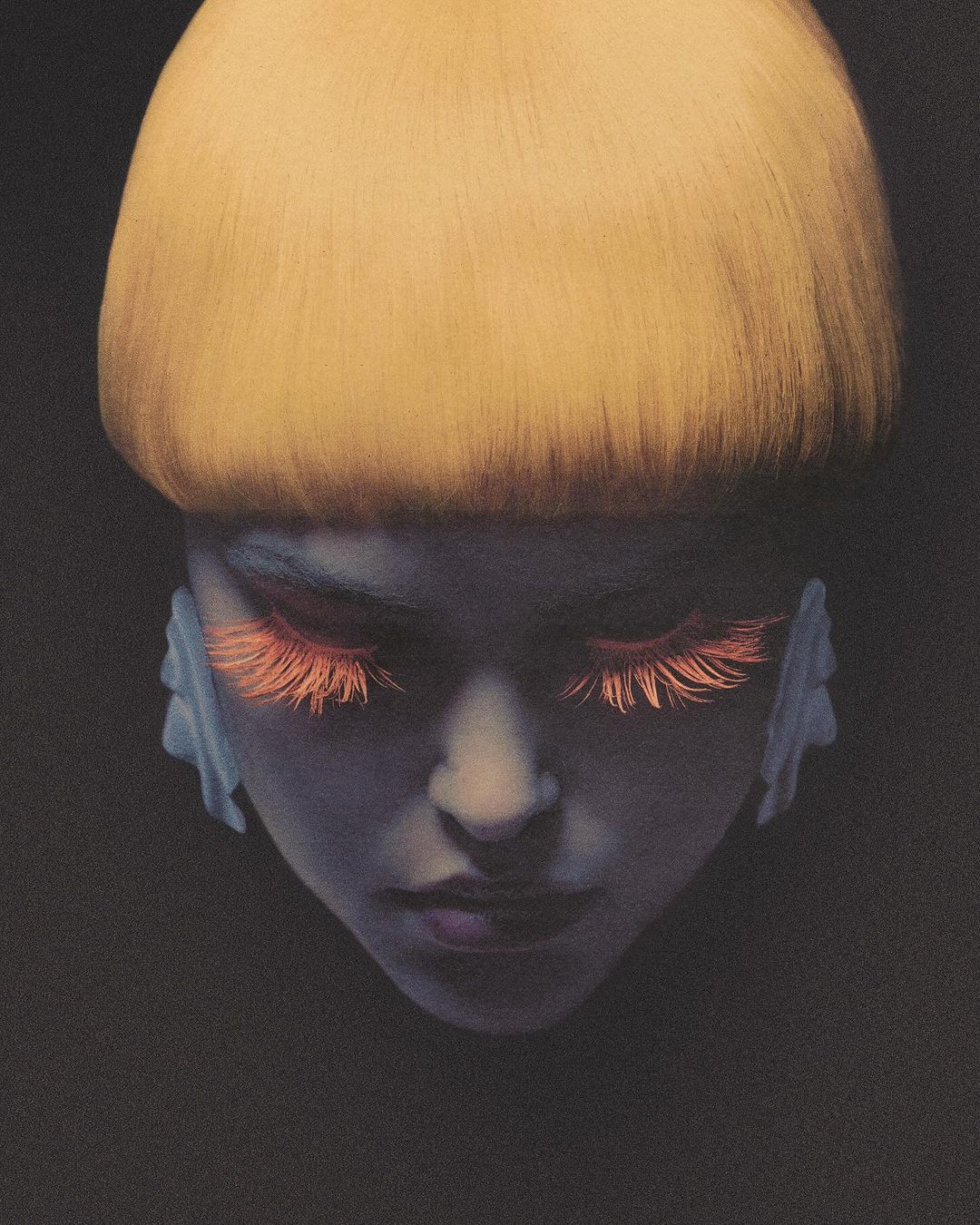 Artistic close-up of a woman's face with vivid orange eyelashes and a blonde bowl cut against a dark background in a portrait.