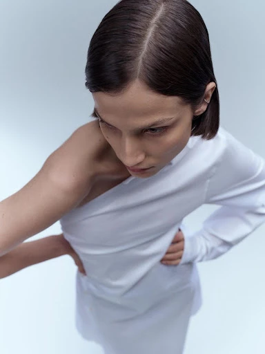 An adult female model in a white asymmetric dress poses in a studio with a neutral backdrop in a fashion shot, shot from above.