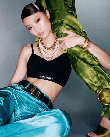 East Asian woman in black bralette and shiny turquoise pants holding a large green satin garment against grey seamless background