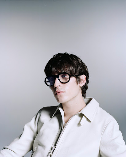 Portrait of a stylish young adult male with dark hair and round black glasses, wearing an off-white jacket, against a grey backdrop.