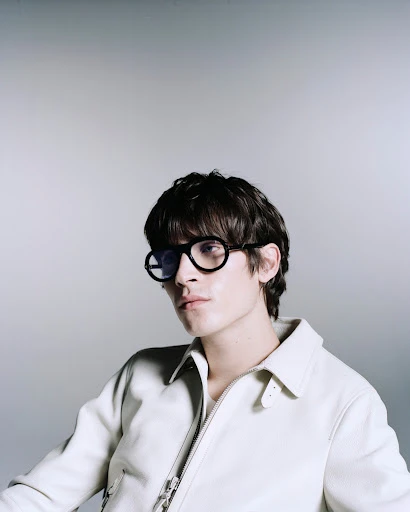 Portrait of a stylish young adult male with dark hair and round black glasses, wearing an off-white jacket, against a grey backdrop.