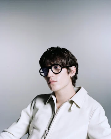Portrait of a stylish young adult male with dark hair and round black glasses, wearing an off-white jacket, against a grey backdrop.