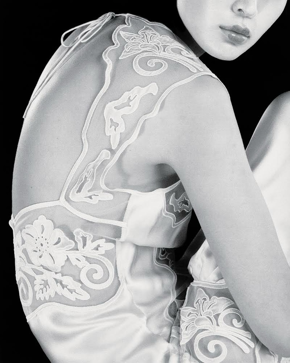 Monochrome portrait of an adult woman with pale skin in a satin dress featuring floral embroidery, posing in a black backdrop.