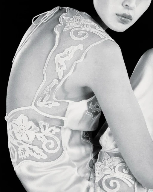 Monochrome portrait of an adult woman with pale skin in a satin dress featuring floral embroidery, posing in a black backdrop.