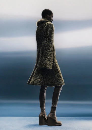 Fashionable adult woman in a green textured coat and matching thigh-high boots poses in a photo studio with a blue background.