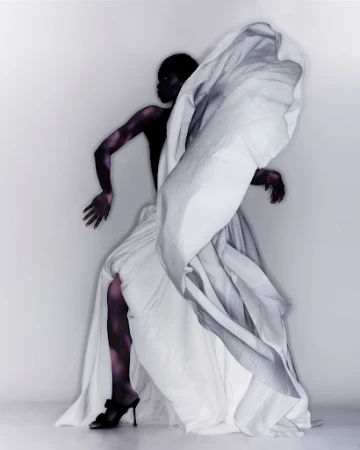Model in a draped white gown and black heels posing against a white seamless background with dramatic lighting.