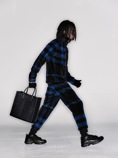 An adult male with dreadlocks strides confidently, carrying a dark tote bag, styled in a blue and black plaid outfit against a neutral backdrop.