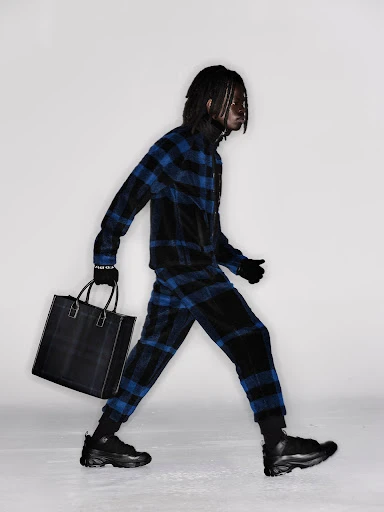 An adult male with dreadlocks strides confidently, carrying a dark tote bag, styled in a blue and black plaid outfit against a neutral backdrop.