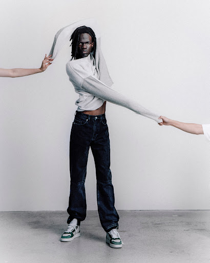An adult male model wearing denim jeans and sneakers poses in a studio environment while partially removing a grey sweater.
