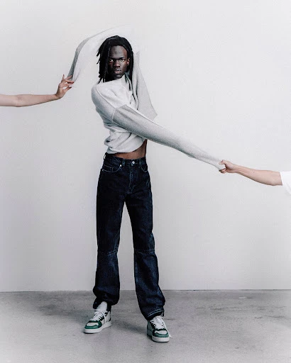An adult male model wearing denim jeans and sneakers poses in a studio environment while partially removing a grey sweater.