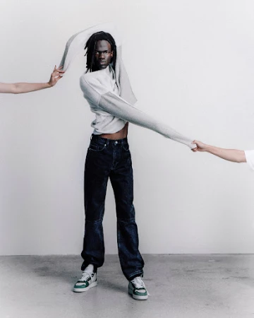 An adult male model wearing denim jeans and sneakers poses in a studio environment while partially removing a grey sweater.