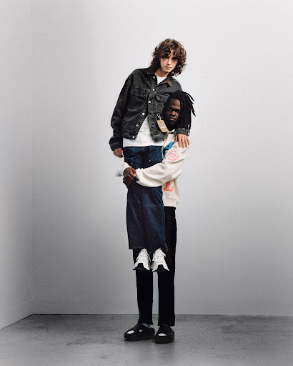 Two men in a minimalist photo studio. One man is giving the other a piggyback ride, both are wearing fashionable casual outfits.