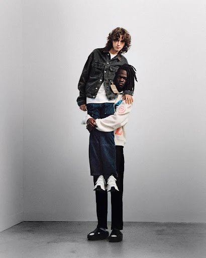 Two men in a minimalist photo studio. One man is giving the other a piggyback ride, both are wearing fashionable casual outfits.