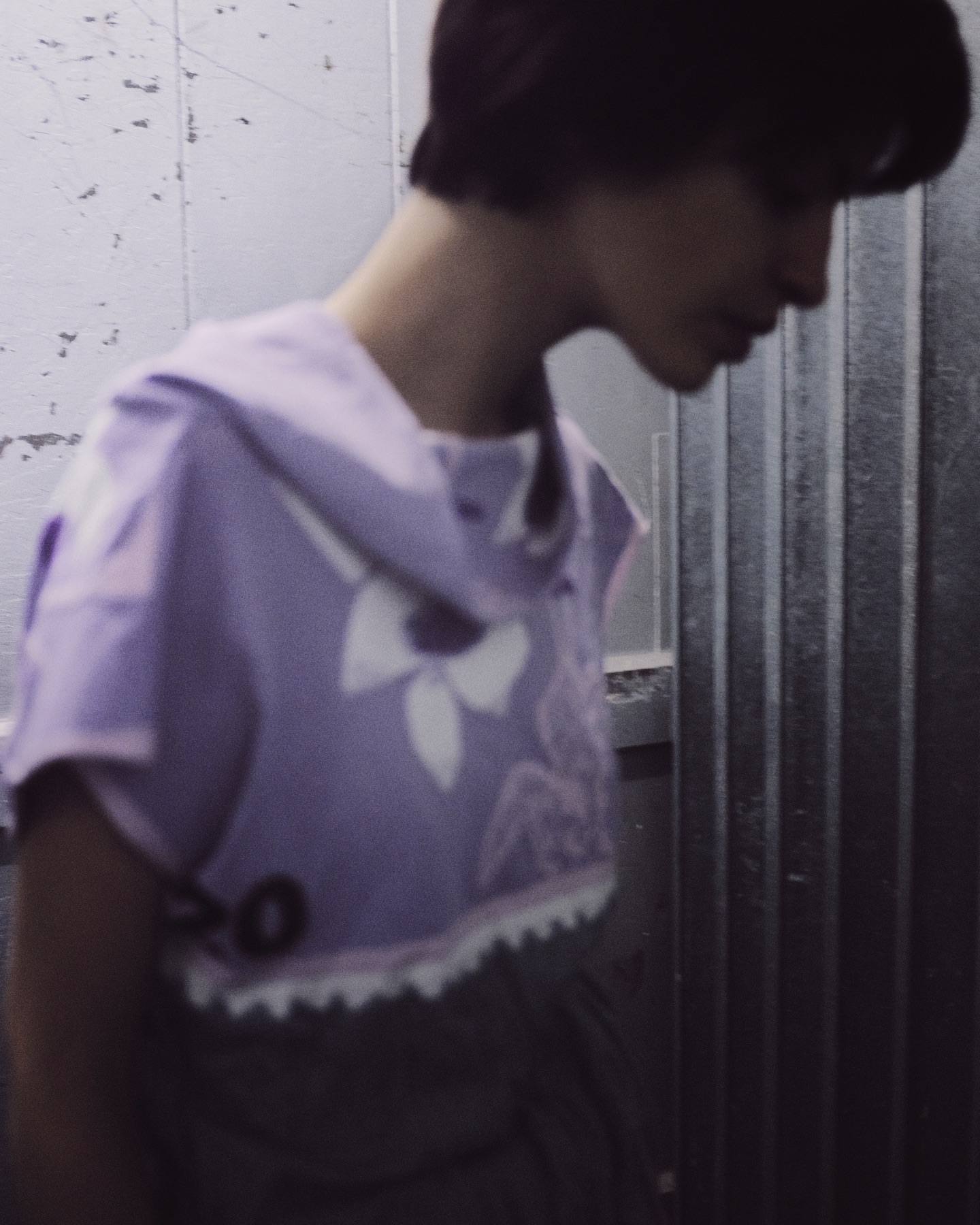 Woman in lavender top poses indoors against a striped gray wall. The top has a white graphic print and lace trim. Soft indoor lighting.