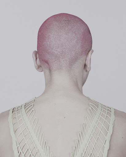 From the back, a person with a pink shaved head wears a net-like, light-green top against a light gray backdrop in a studio setting.
