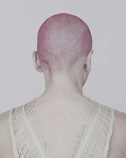 From the back, a person with a pink shaved head wears a net-like, light-green top against a light gray backdrop in a studio setting.