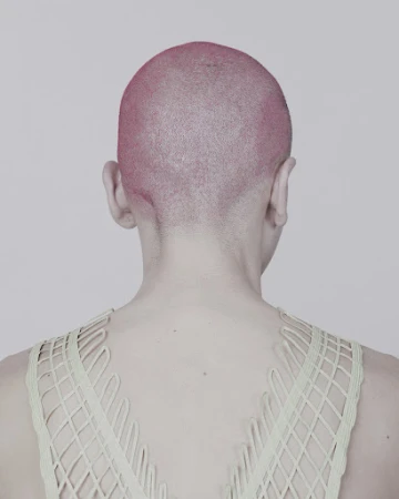 From the back, a person with a pink shaved head wears a net-like, light-green top against a light gray backdrop in a studio setting.