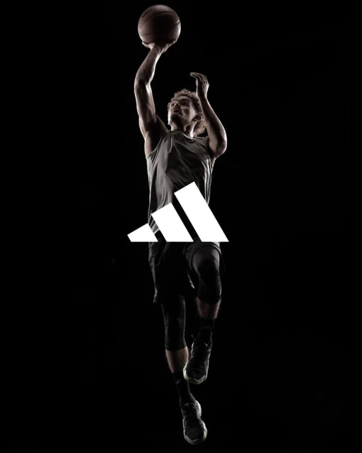 Silhouette of male basketball player in mid-air shooting basketball with Adidas logo on dark background in athletic sportswear.