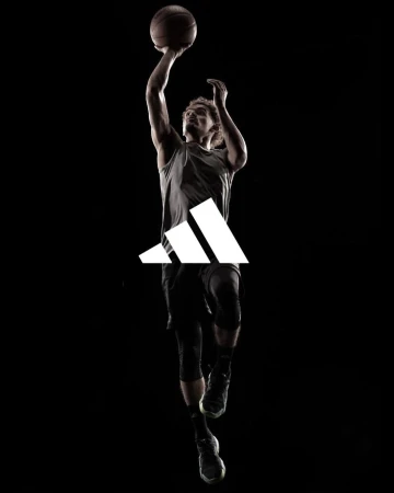 Silhouette of male basketball player in mid-air shooting basketball with Adidas logo on dark background in athletic sportswear.