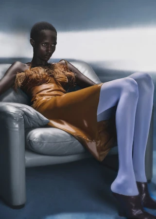 Black model with short hair wearing a feathered brown dress and blue tights seated in a grey leather armchair.