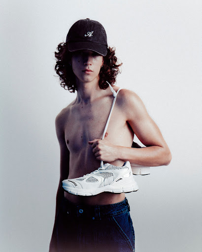Studio shot of a shirtless adult male wearing a baseball cap and jeans, holding a white sneaker against a plain background.
